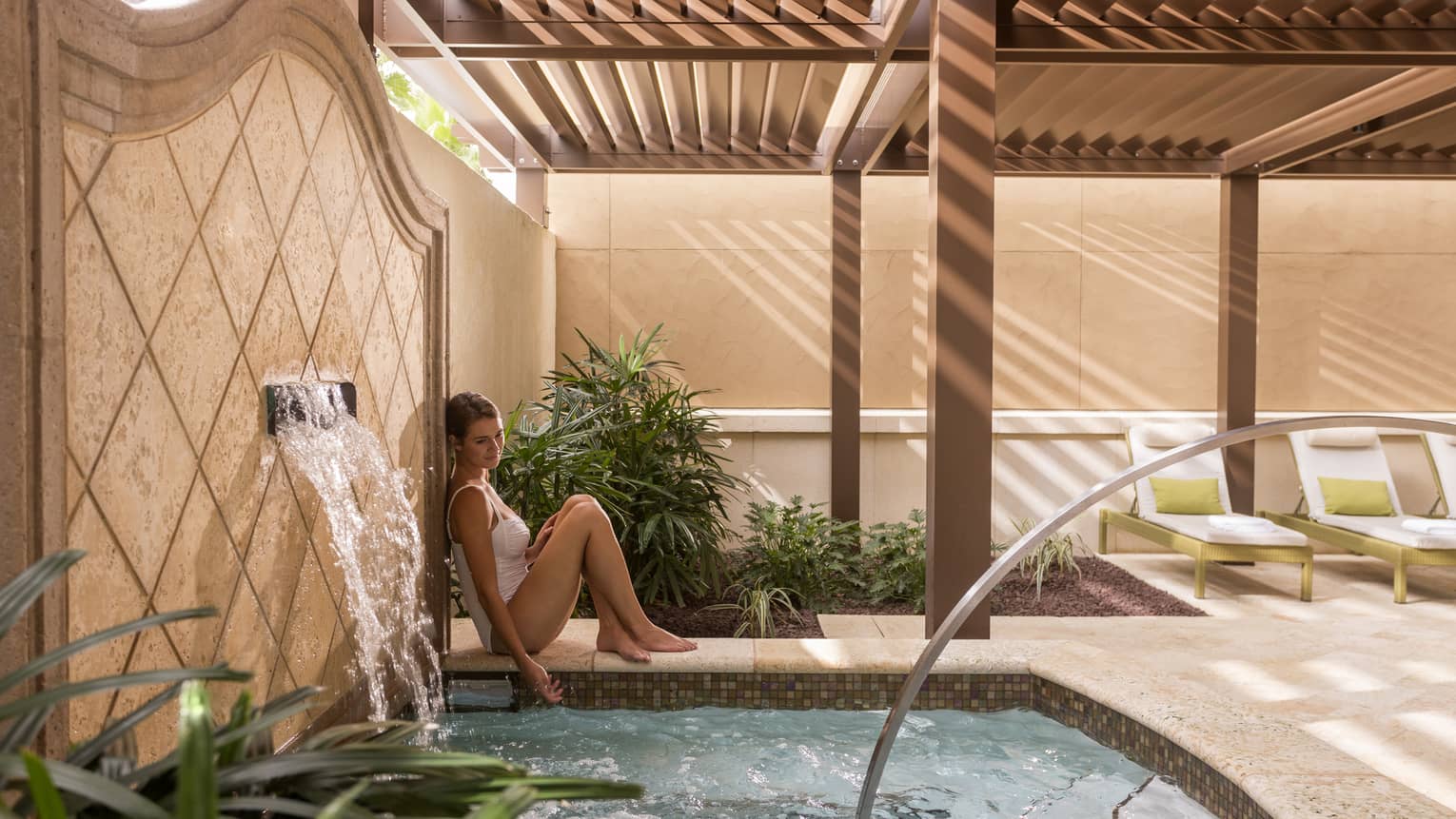 Woman wearing swimsuit leans against tile wall with fountain by small spa pool