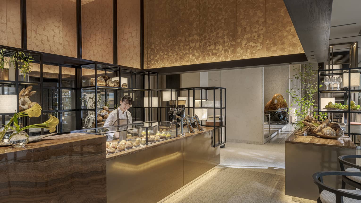 A person stands behind a counter displaying food items in an open, well-lit space decorated in warm neutral tones; the space also includes chairs, open shelves, an additional counter and a freestanding island