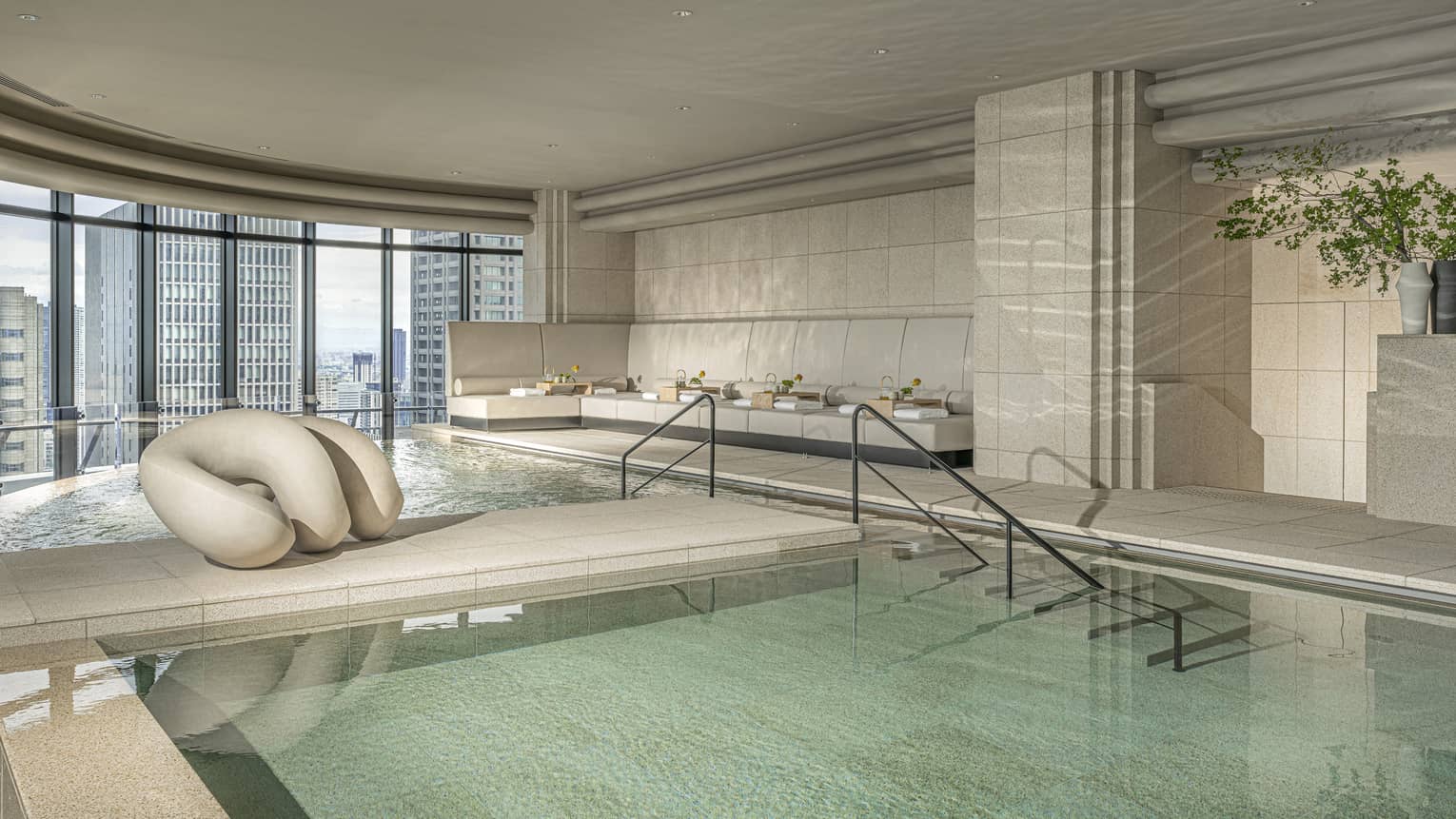 An indoor pool area with steps and rails leading into the pools in an open space with beige walls, floor and ceiling and a wall of large windows letting in light and city views