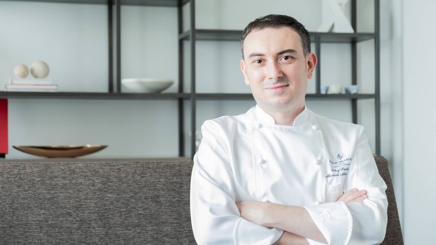 Smiling man wearing white chef's coat and sitting on couch poses for camera with arms crossed