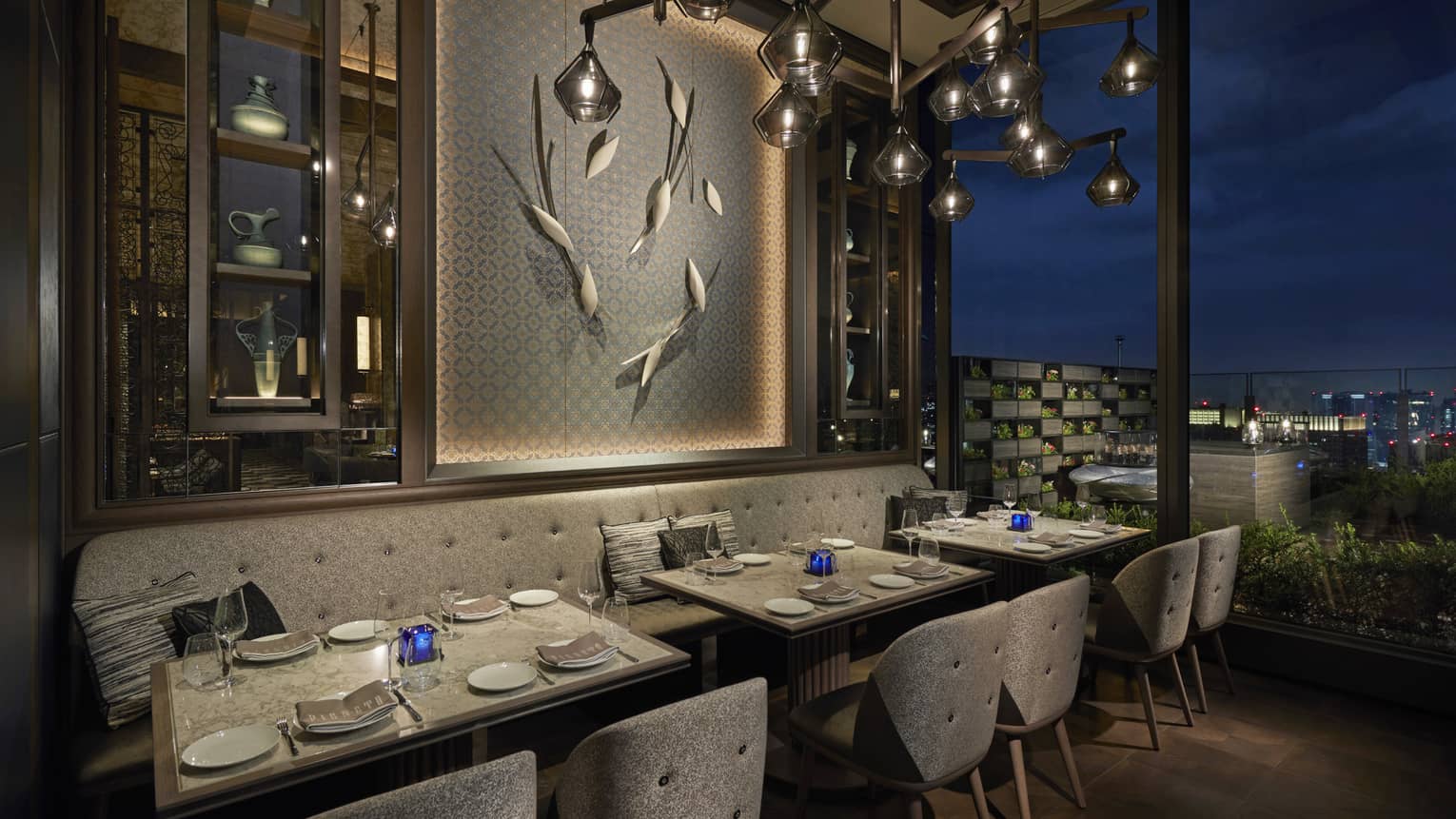 Tables set for two overlook nighttime skyline, floor-to-ceiling windows, intimate lighting, grey colour scheme 