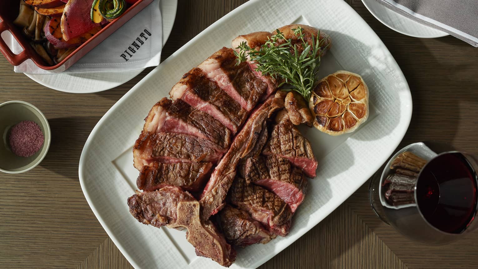 Aerieal view of medium-rare sliced T-bone steak with rosemary and roasted garlic garnishes on rectangular white plate, glass of red wine nearby