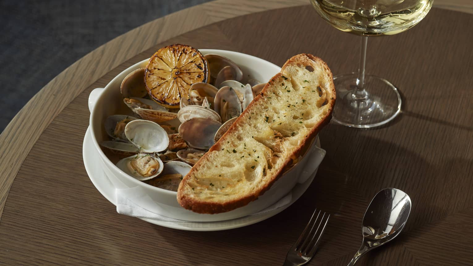 Closeup shot of whole clams in white Corningware bowl, garnished with charred lemon half and rustic slice of bread, glass of white whine nearby