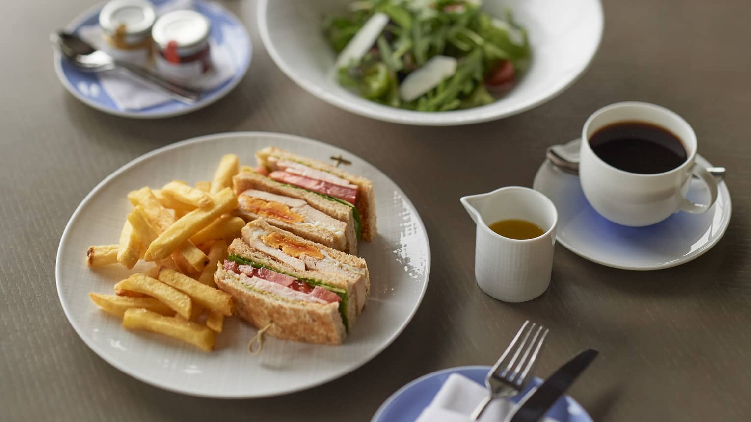 Club sandwich and French fries on round white plate, side salad in matching white bowl, mug of black coffee