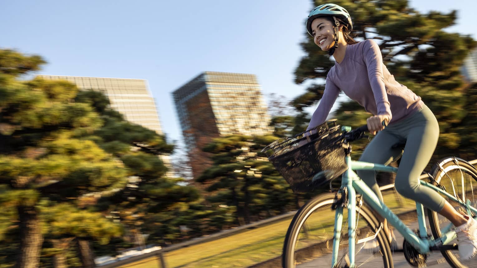 Smiling woman on teal cruiser bike with matching helmet rides by mature trees and skyscrapers of downtown Tokyo