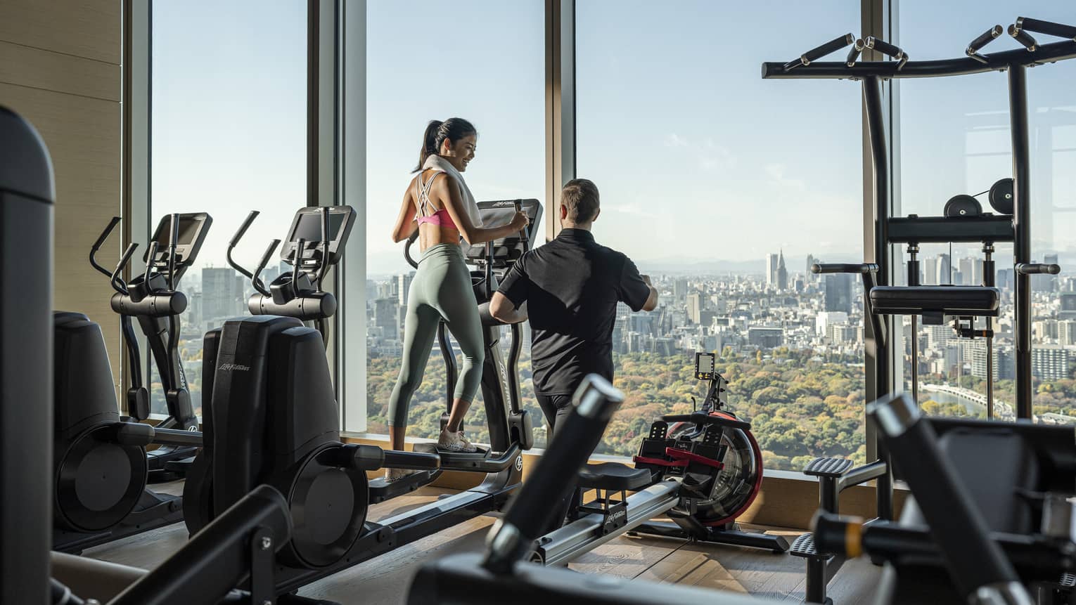 Woman trains on treadmill with male trainer, overlooking downtown Tokyo
