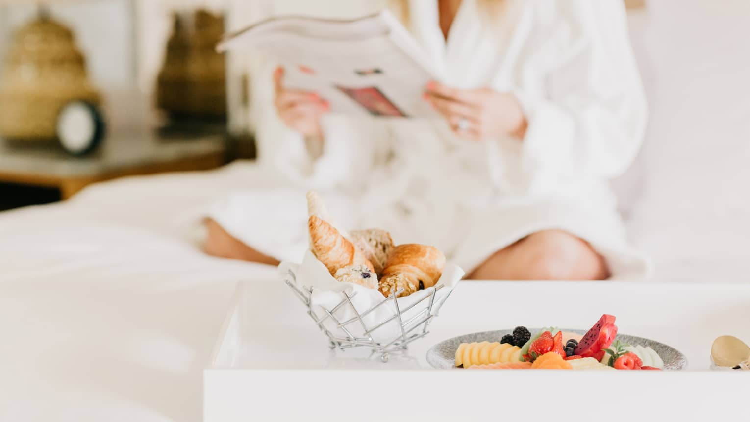 Pastries, fruit on in room dining tray, woman wearing white bath robe reads in background