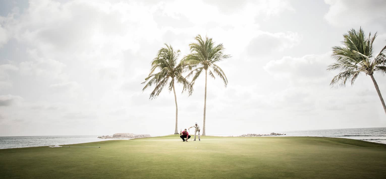 Two men kneel on golf green under two palm trees, ocean on each side