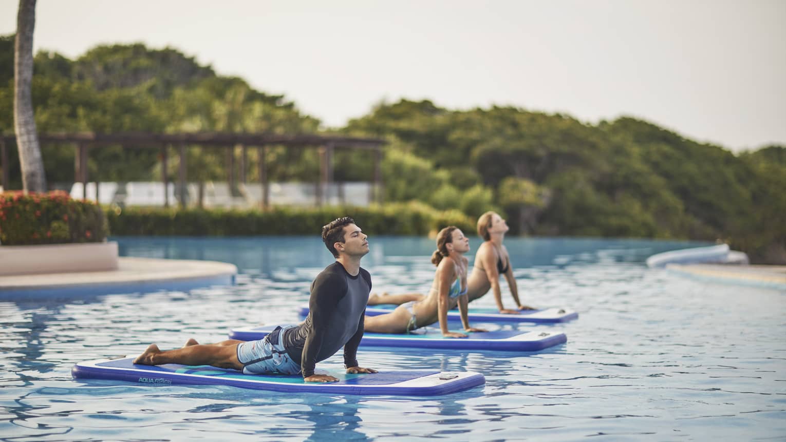 Man, two women in yoga poses on paddle boards on swimming pool