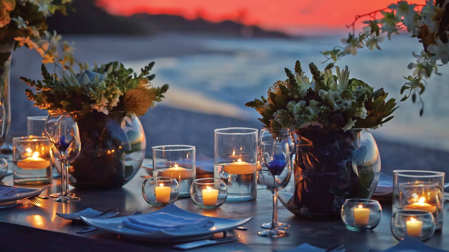 Large round vases of flowers and many small candles in glass holders grace a table facing the beach under a pink-orange sky. 