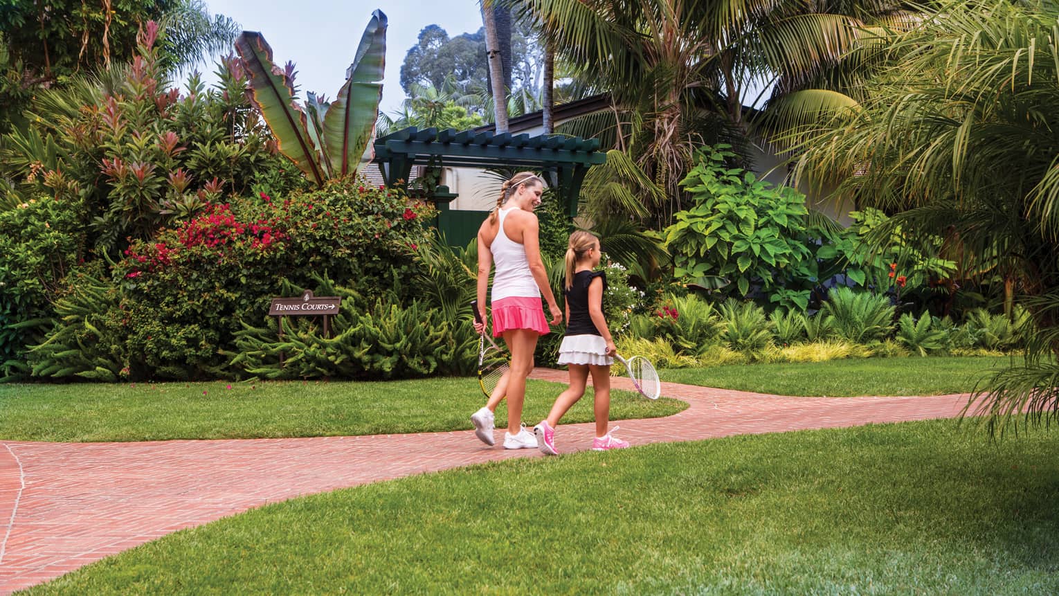 Woman and young girl holding tennis rackets walk down brick path, tropical garden