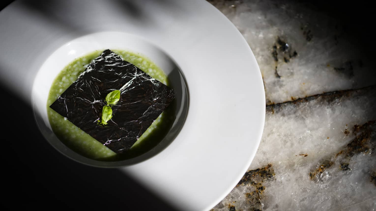 Black Squid Ink Square atop Green Risotto in white soup bowl on marble counter