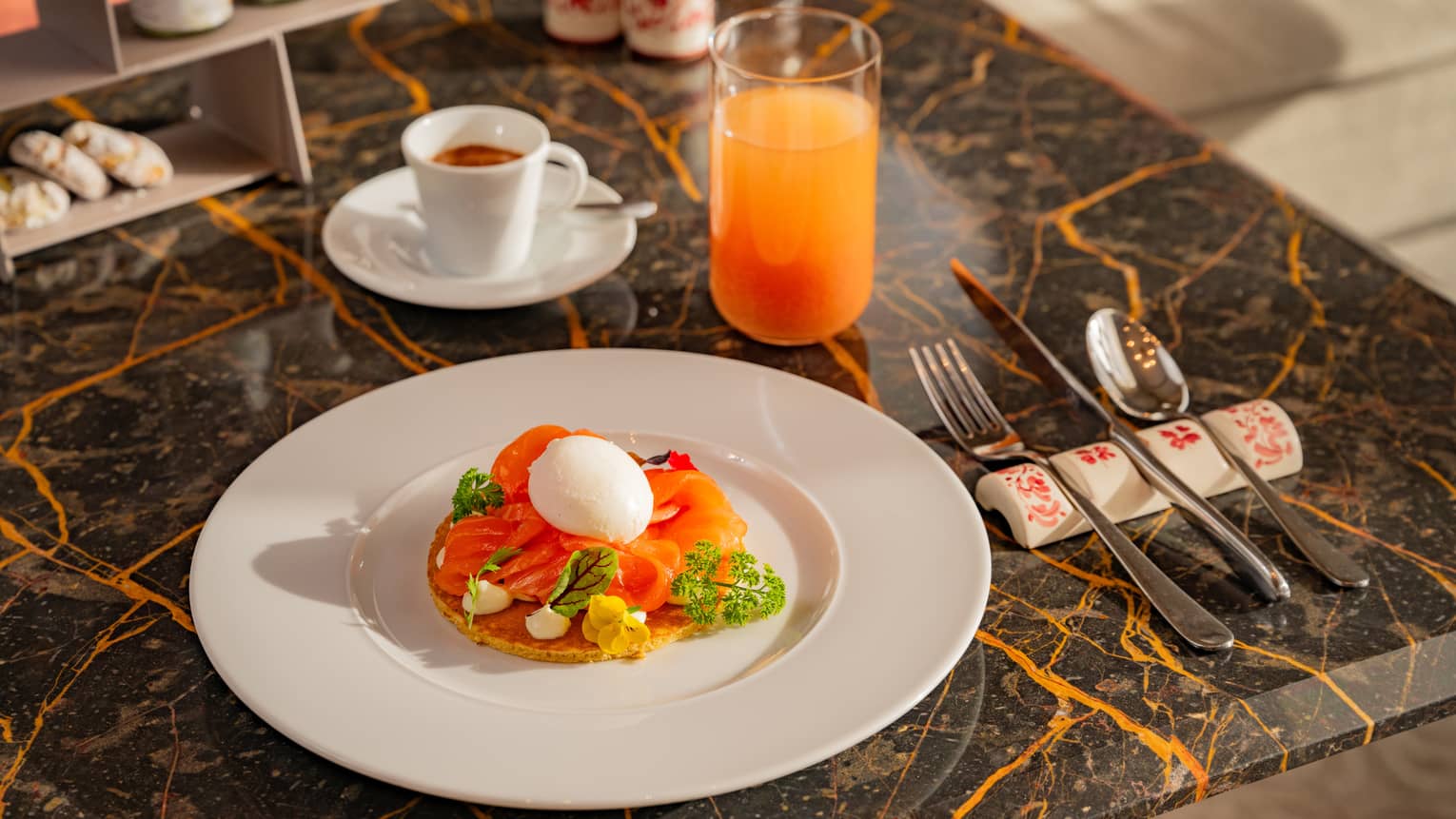 Pancake topped with sliced salmon and hard-boiled egg beside flatware, cup of espresso and glass of juice