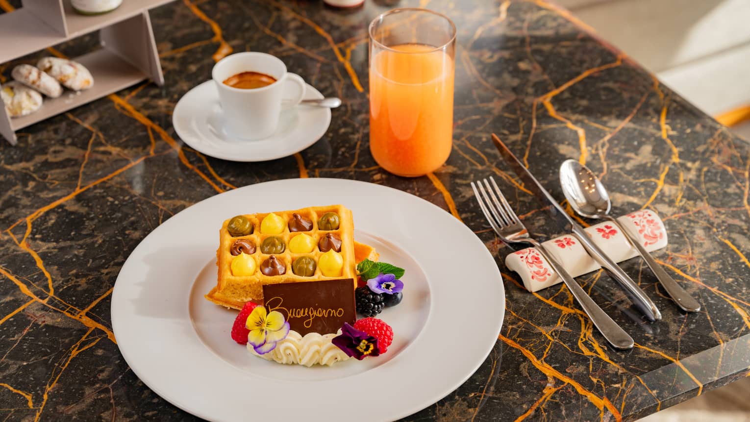 Waffle filled with cream and garnished with chocolate, pansies and mixed berries on white plate beside flatware, cup of espresso and glass of juice