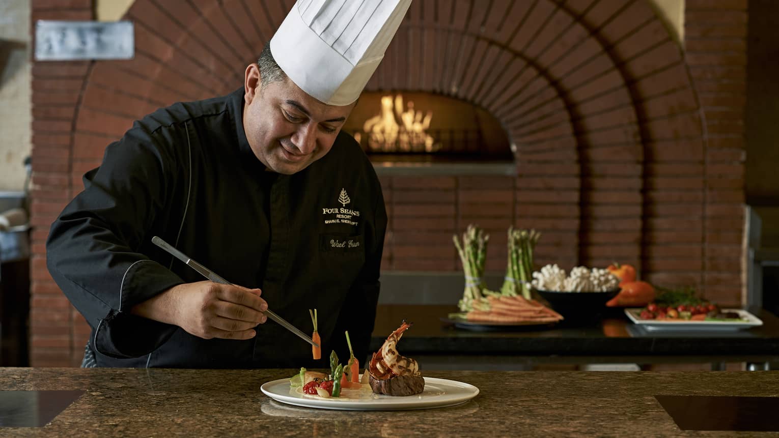 Chef in uniform holds tweezers, arranges vegetables on plate with large steak, lobster tail by wood-burning oven