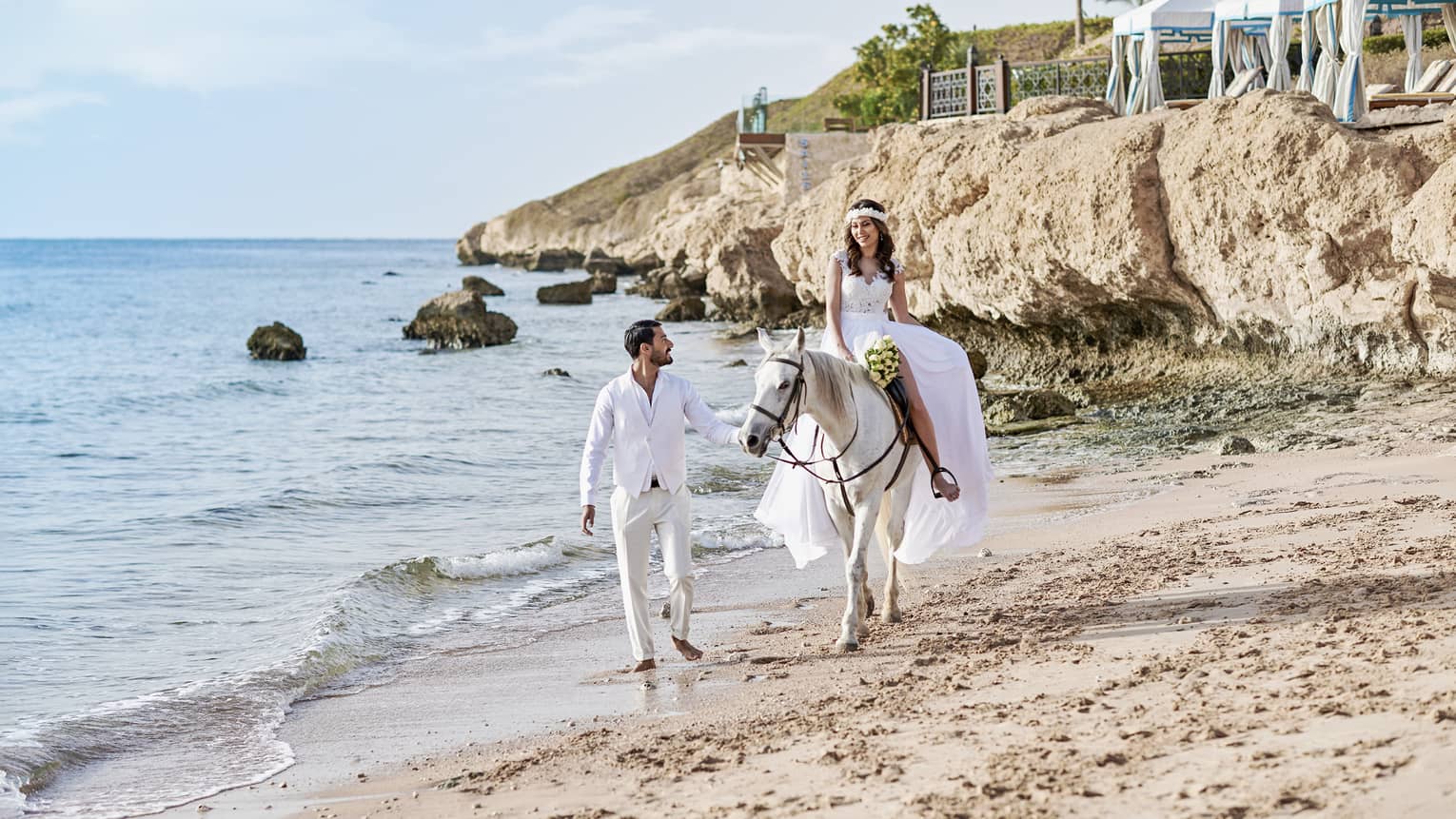 Groom leads bride riding white horse across sand beach