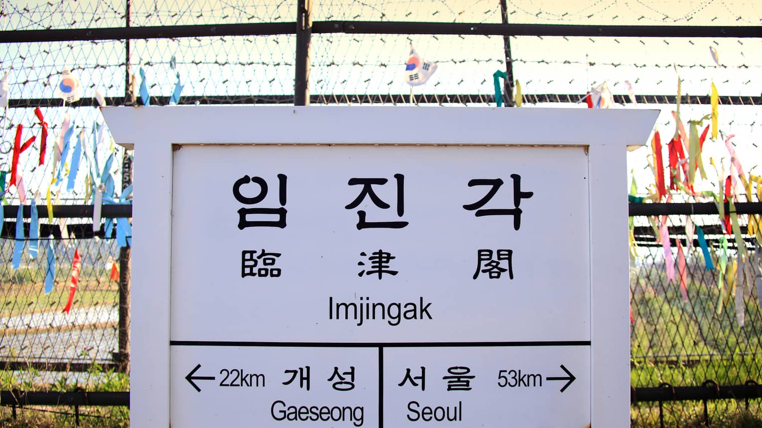 Korean and English text on white sign in front of barbwire fence with ribbons, marking border between North and South Korea.