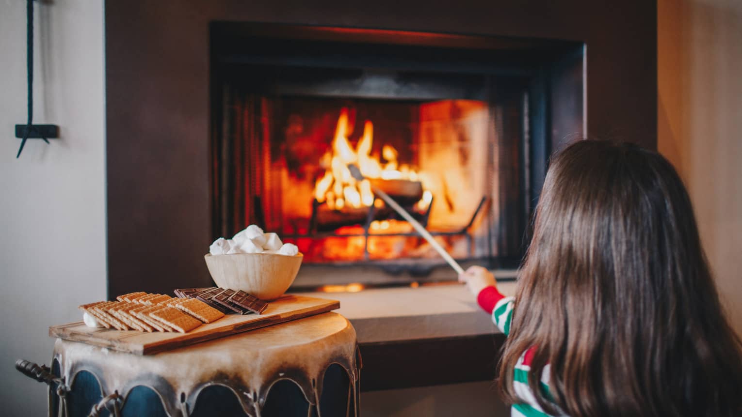 A little girl in red and green pajamas roasts marshmallows in a fireplace