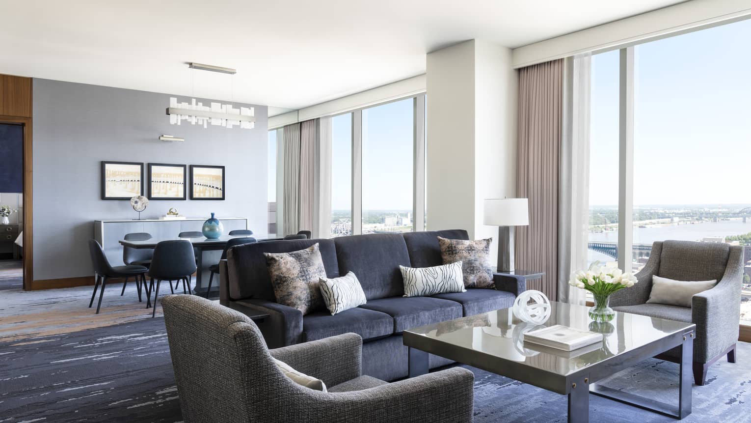 Hotel suite living room with dark blue sofa, two grey arm chairs, large square coffee table and wall of windows