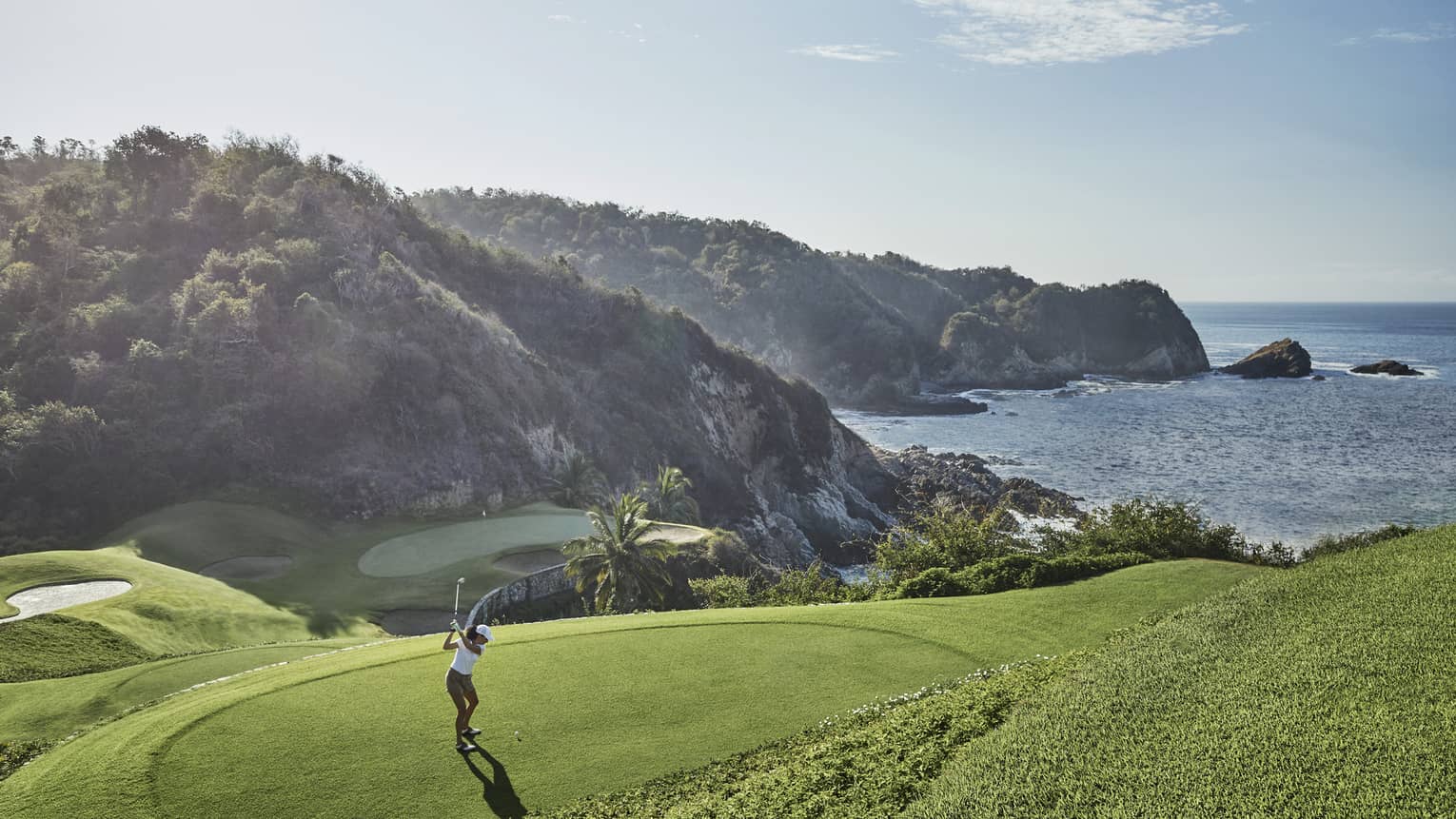 Golfer in mid-backswing, ready to strike toward a distant green at the base of an immense forested hill bordering the ocean. 