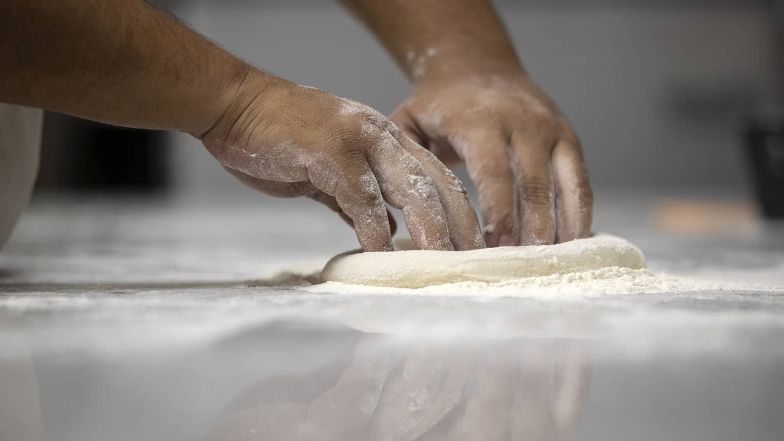 Flour dusted hands shape a small round of pizza dough on a flour-covered stainless steel counter. 