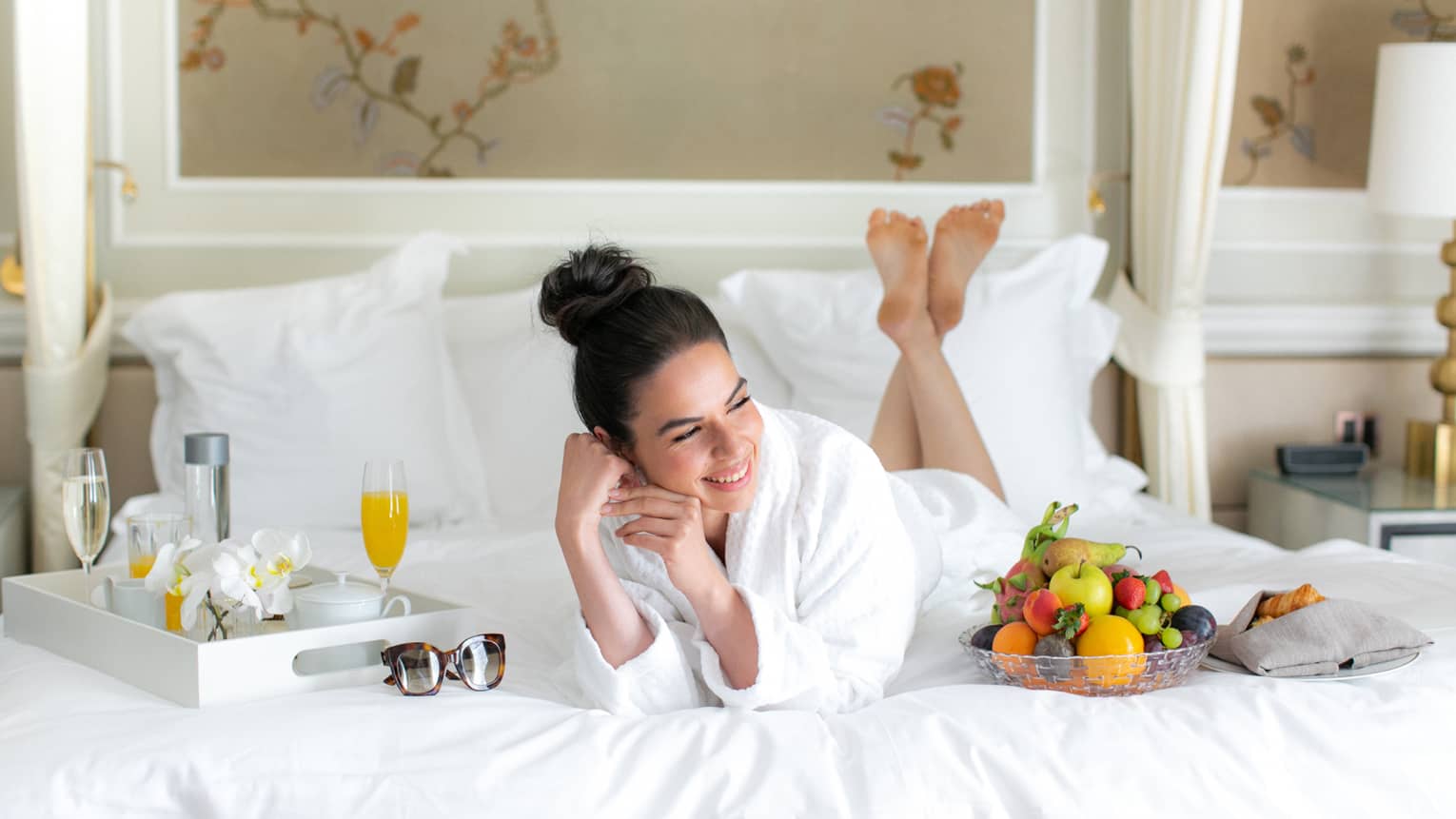 A woman in a spa robe laying on a bed with juice and fruit.