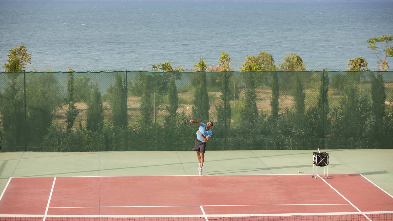 Man in blue shirt and black shorts serving a tennis ball on a seafront tennis court