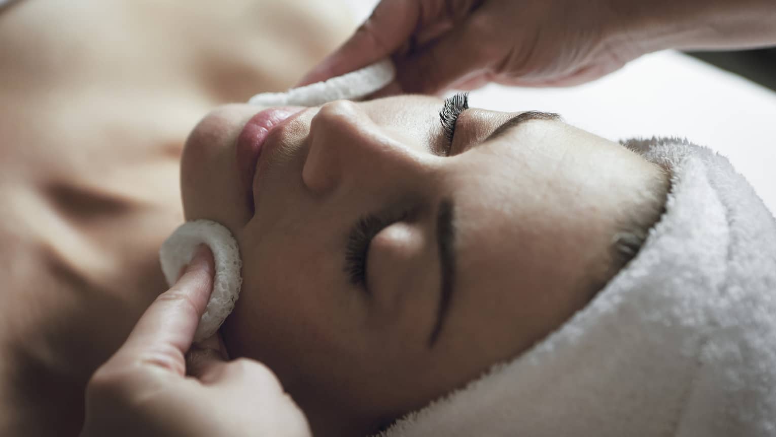 A detail of hands rubbing a sponge on a woman's face as she lies on a massage table with her eyes closed and hair wrapped up in a white towel
