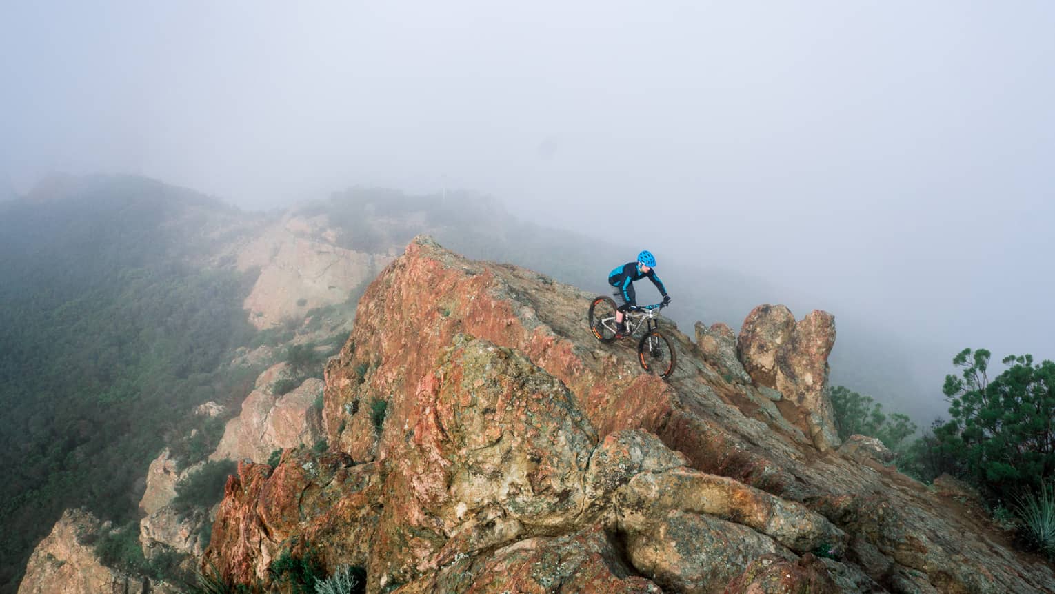 Cyclist rides down large boulder at top of misty mountain