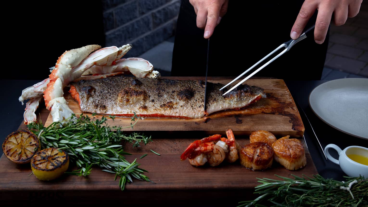 Closeup of chef preparing to carve a side of steelhead trout on a cedar plank, rosemary sprigs and charred lemon garnishes around