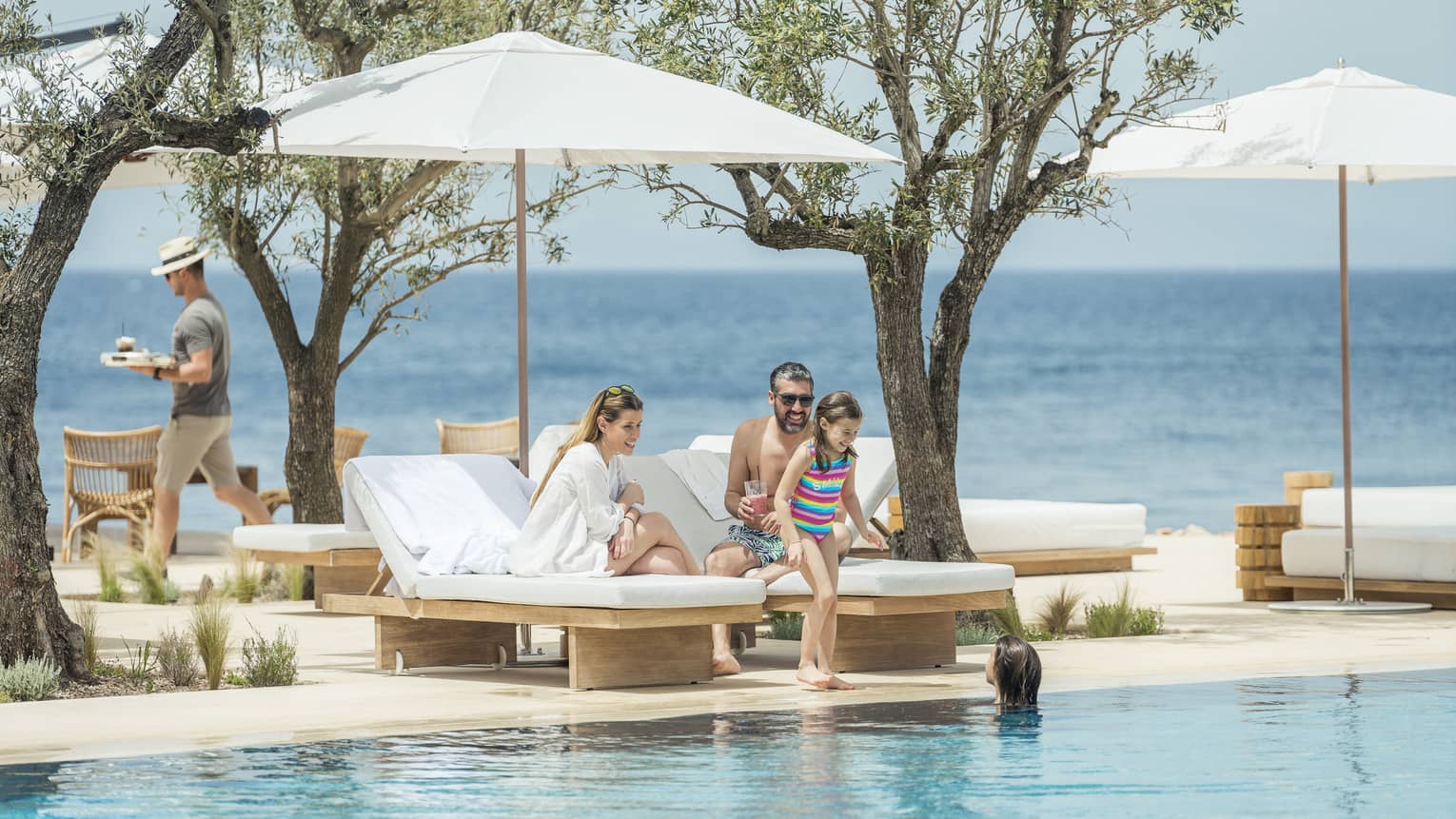 A man and woman sit on white lounge chairs while their two kids play in the pool overlooking the ocean