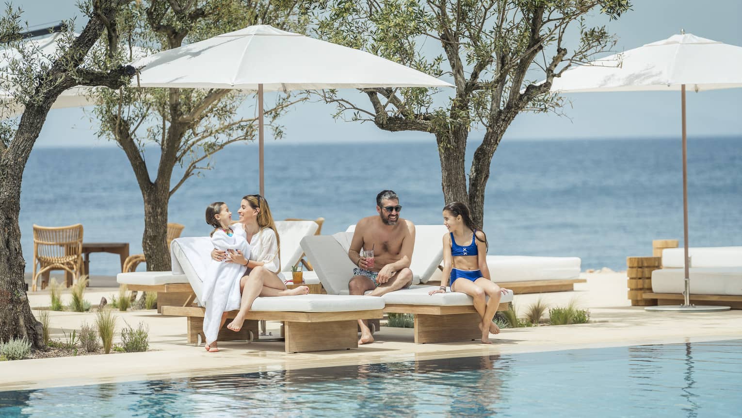 A family of four mingles on two white lounge chairs next to a pool, white umbrellas, sea in the distance