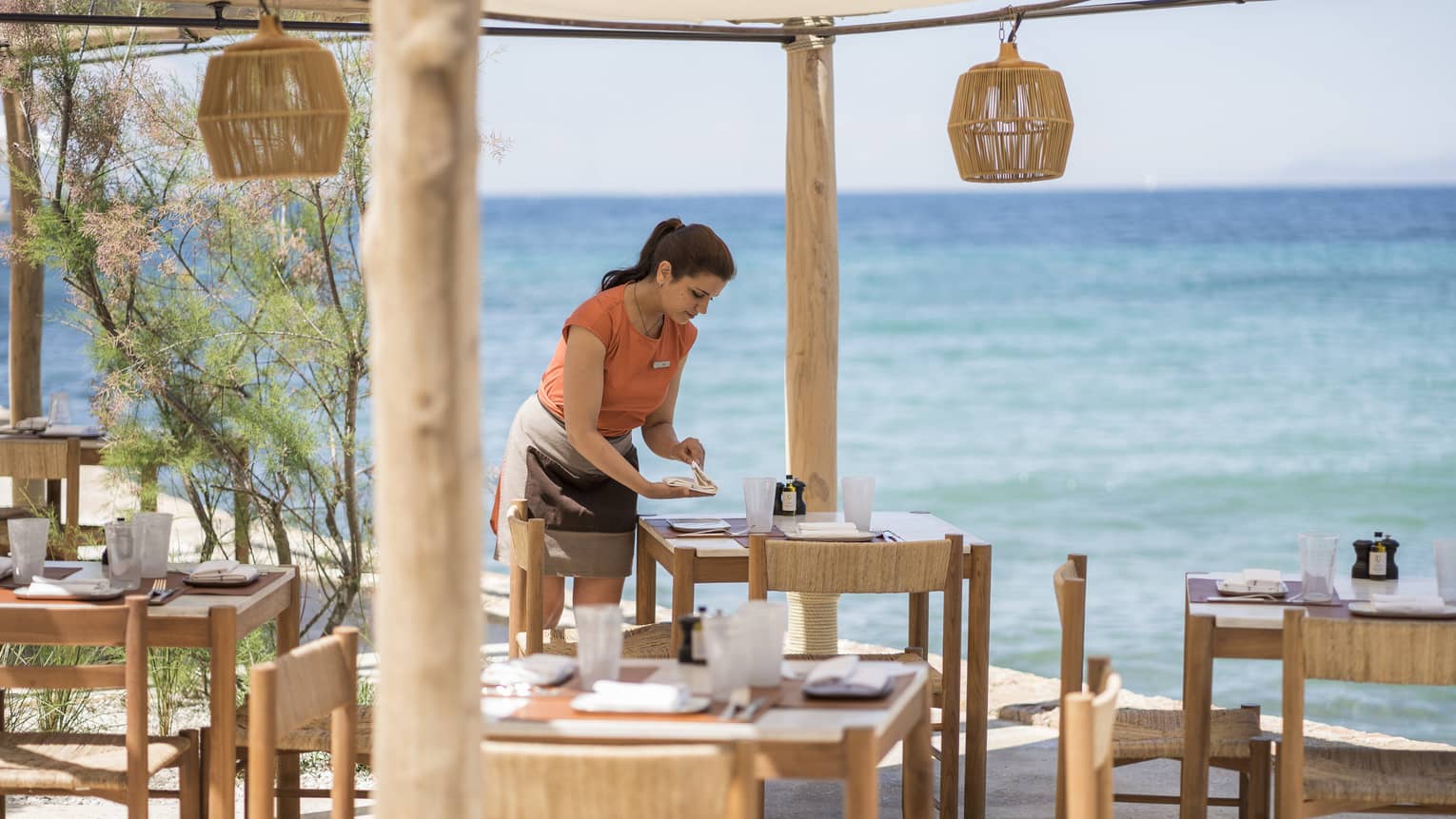 Woman in orange top sets wooden table at outdoor Taverna with wooden lanterns by ocean