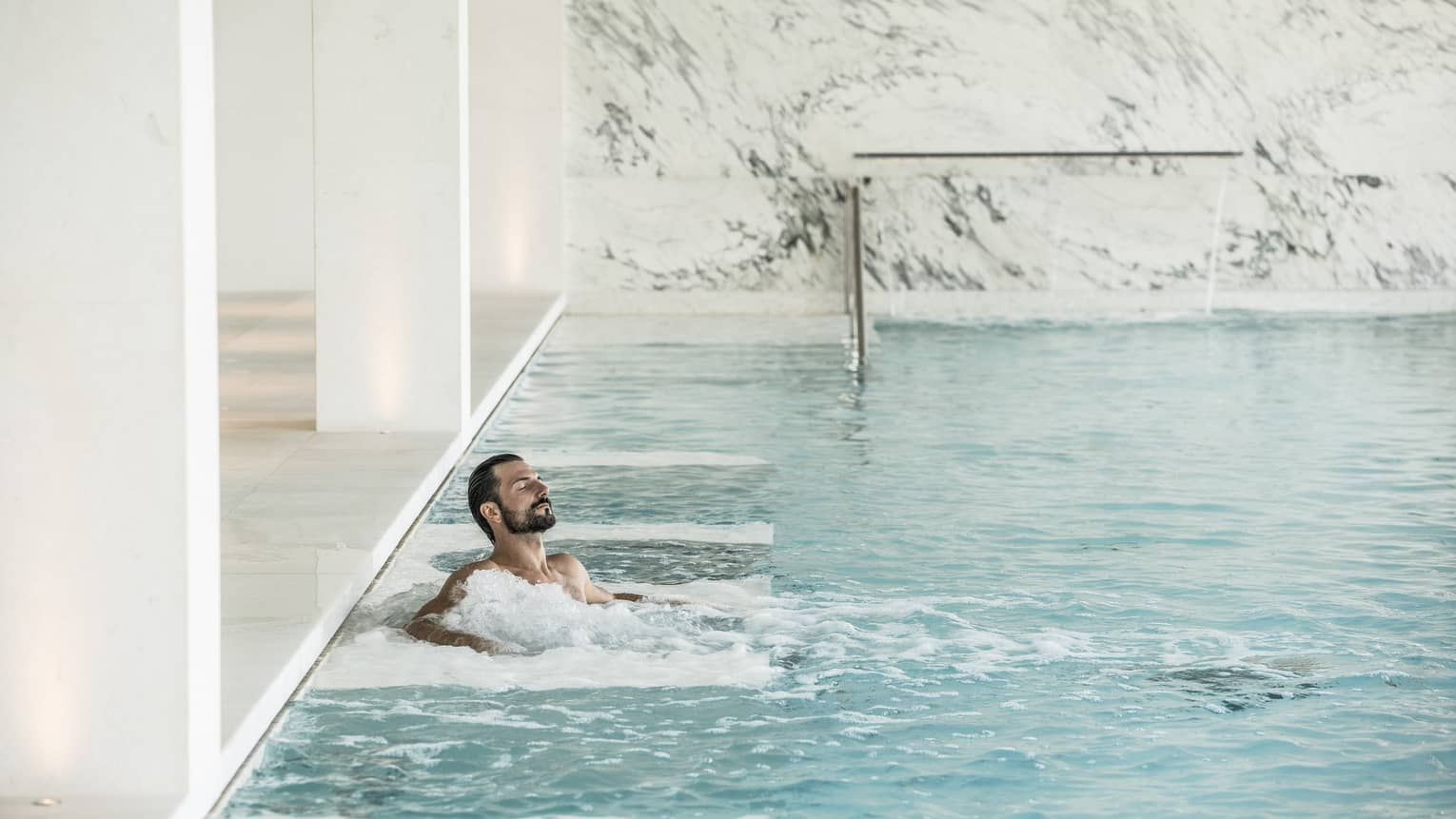 Man with beard relaxes in spa waters, surrounded by white marble