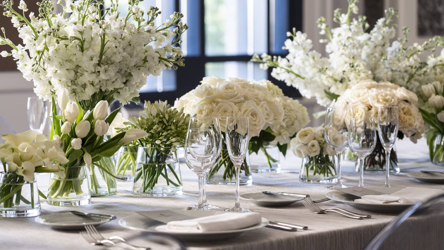A grey table with an assortment of white flowers in a row, along with white plates and wine glasses.