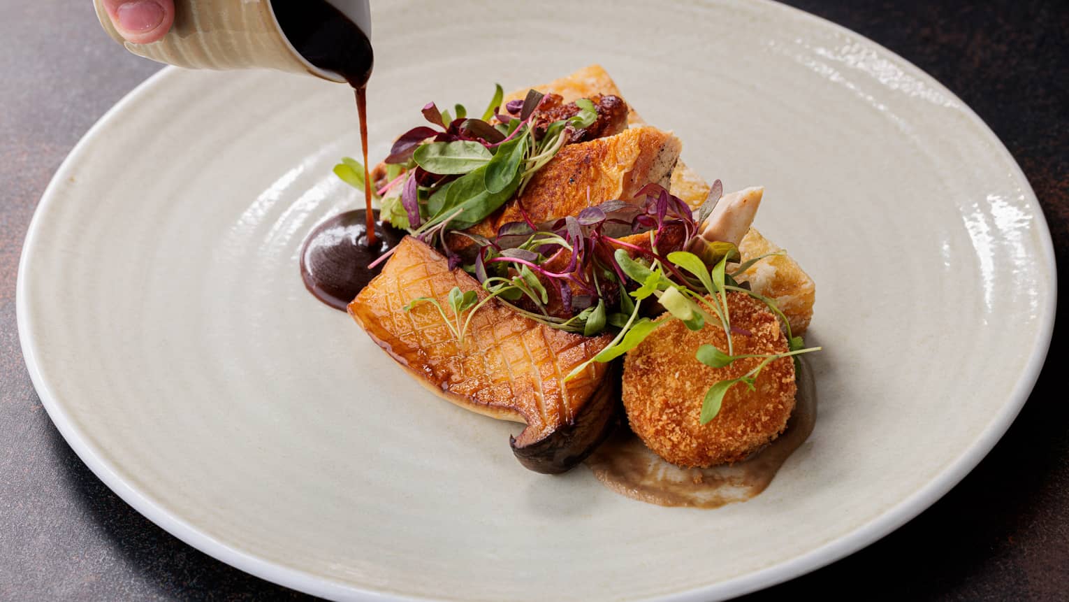 Pan-seared chicken breast with a zuchini ribbon, confit tomato, shallots, corn puree on a white plate, with a natural jus being poured on gently