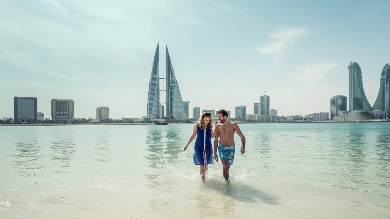 A man and woman run arm in arm through the shallow ocean water with Bahrain Bay skyscrapers in the background