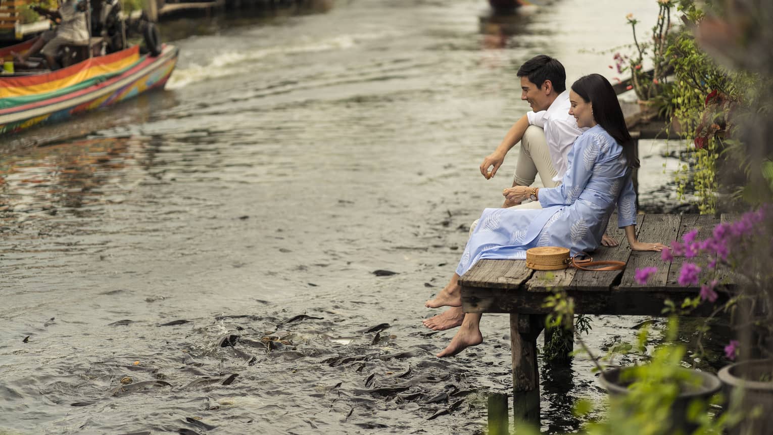 Couple sits on the dock of the Chao Phraya River, hungry fish swarming beneath their dangling feet