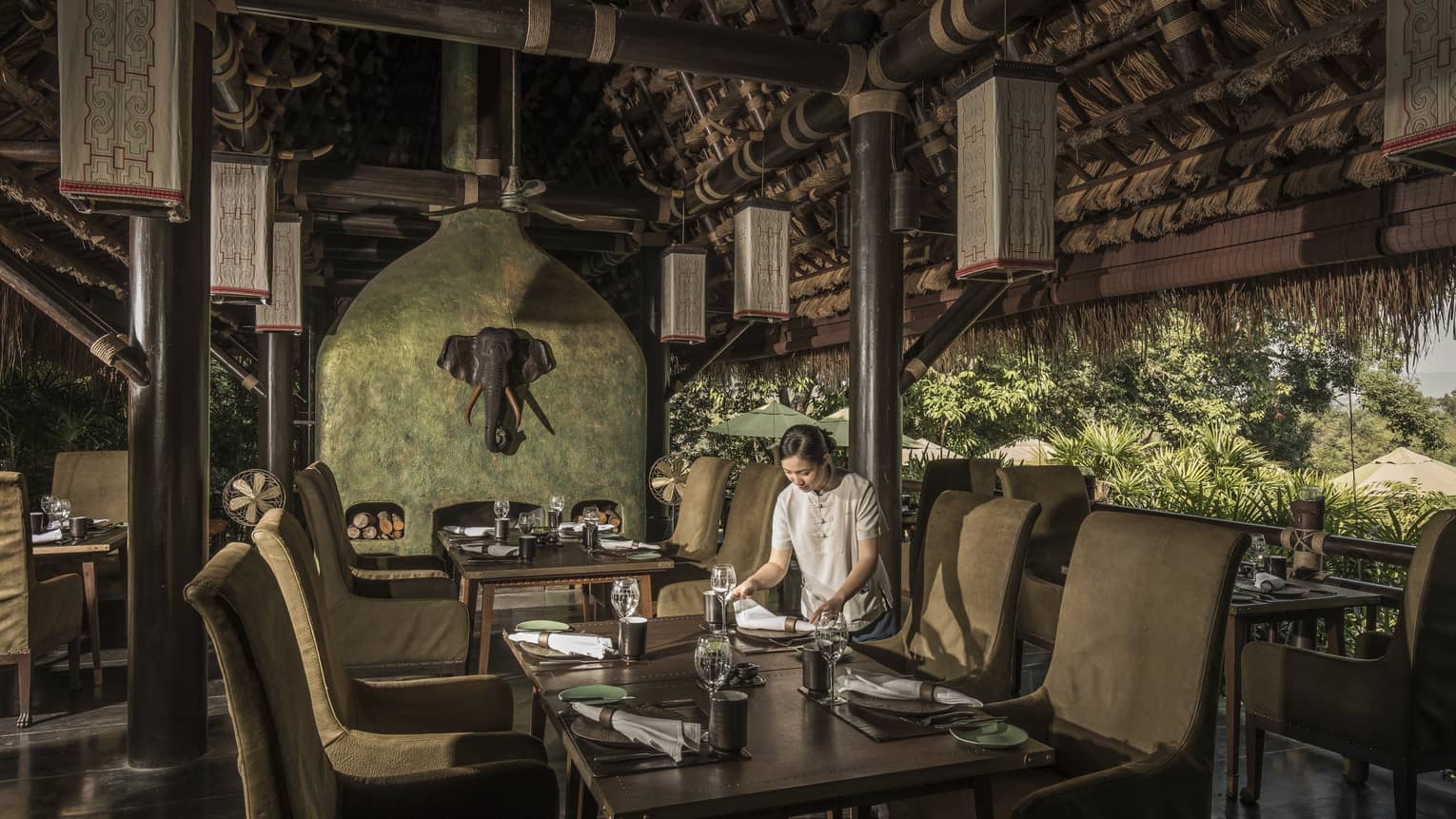 A woman sets the table in an open-air restaurant, natural-coloured furnishings, elephant head decoration on wall