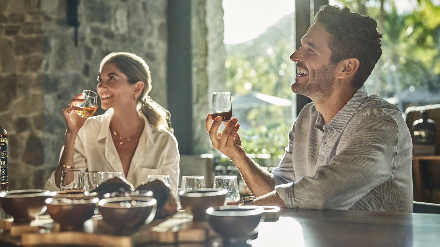 A man and a woman sit at a counter looking up and smiling and each holding a glass of rum with various other glasses and ingredients sit in front of them on the counter