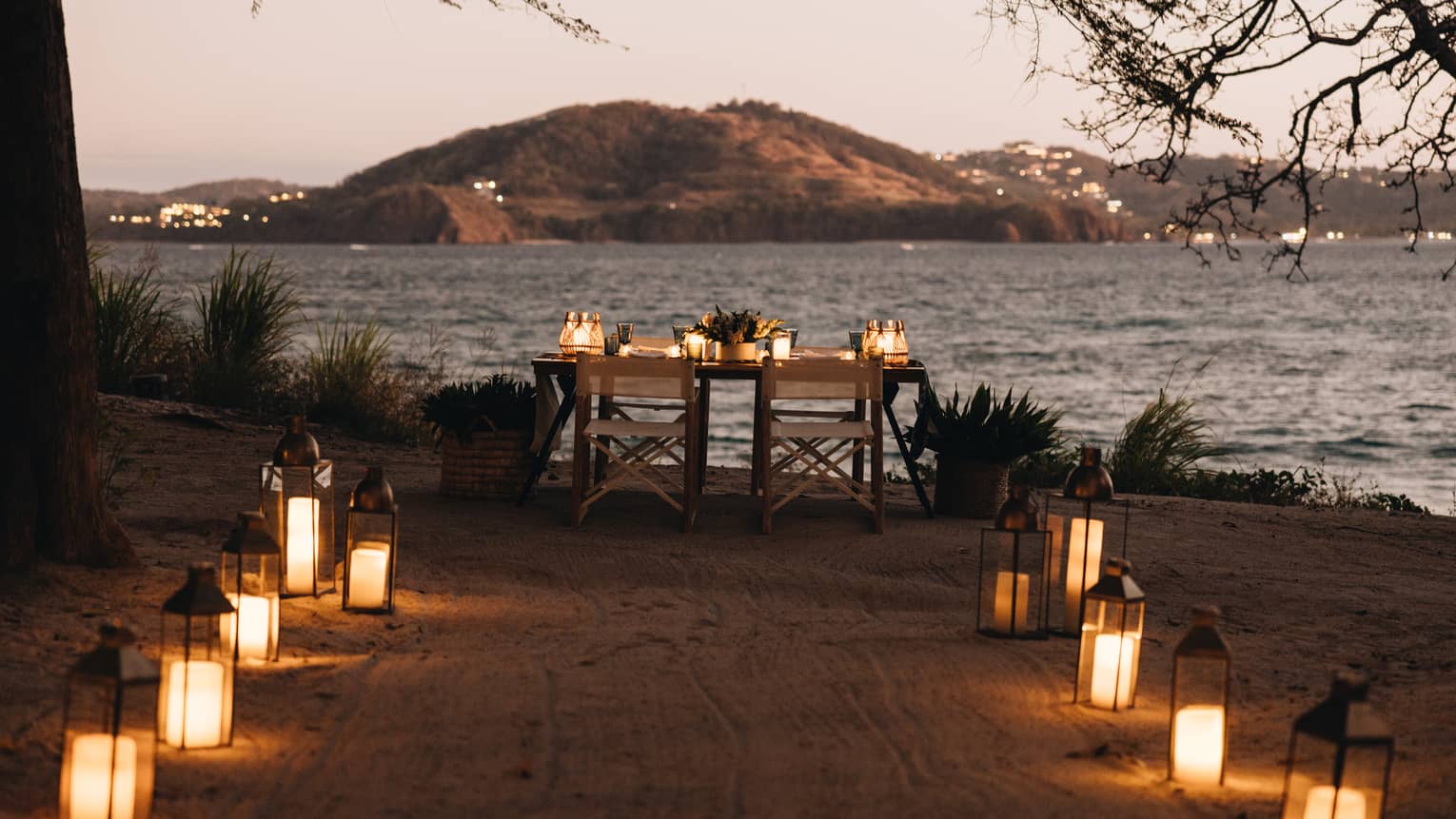 Lanterns line a path on the sand leading up to a table set for two overlooking the ocean