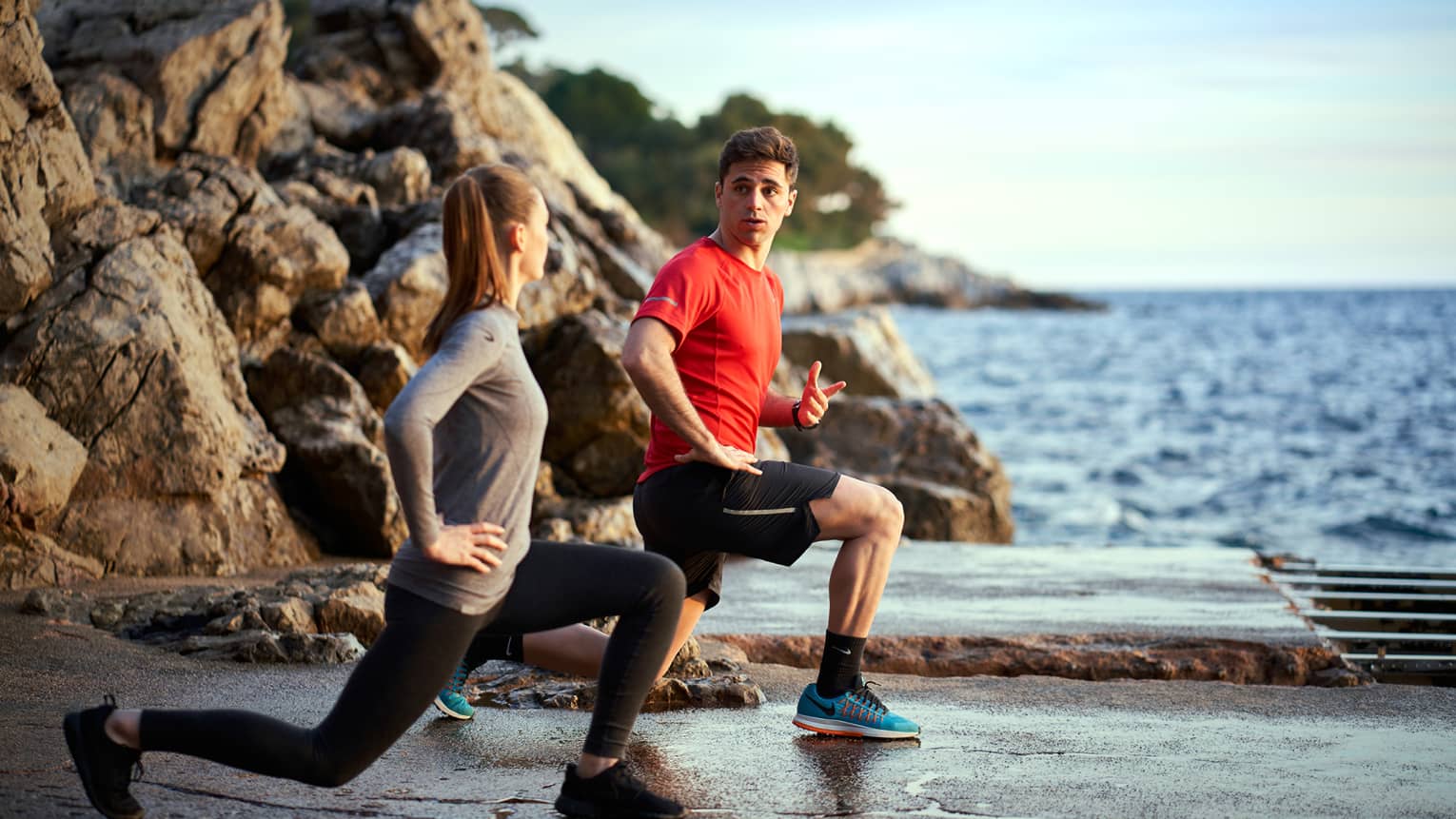 A man trains a woman while doing lunges outside next to the sea and rocks