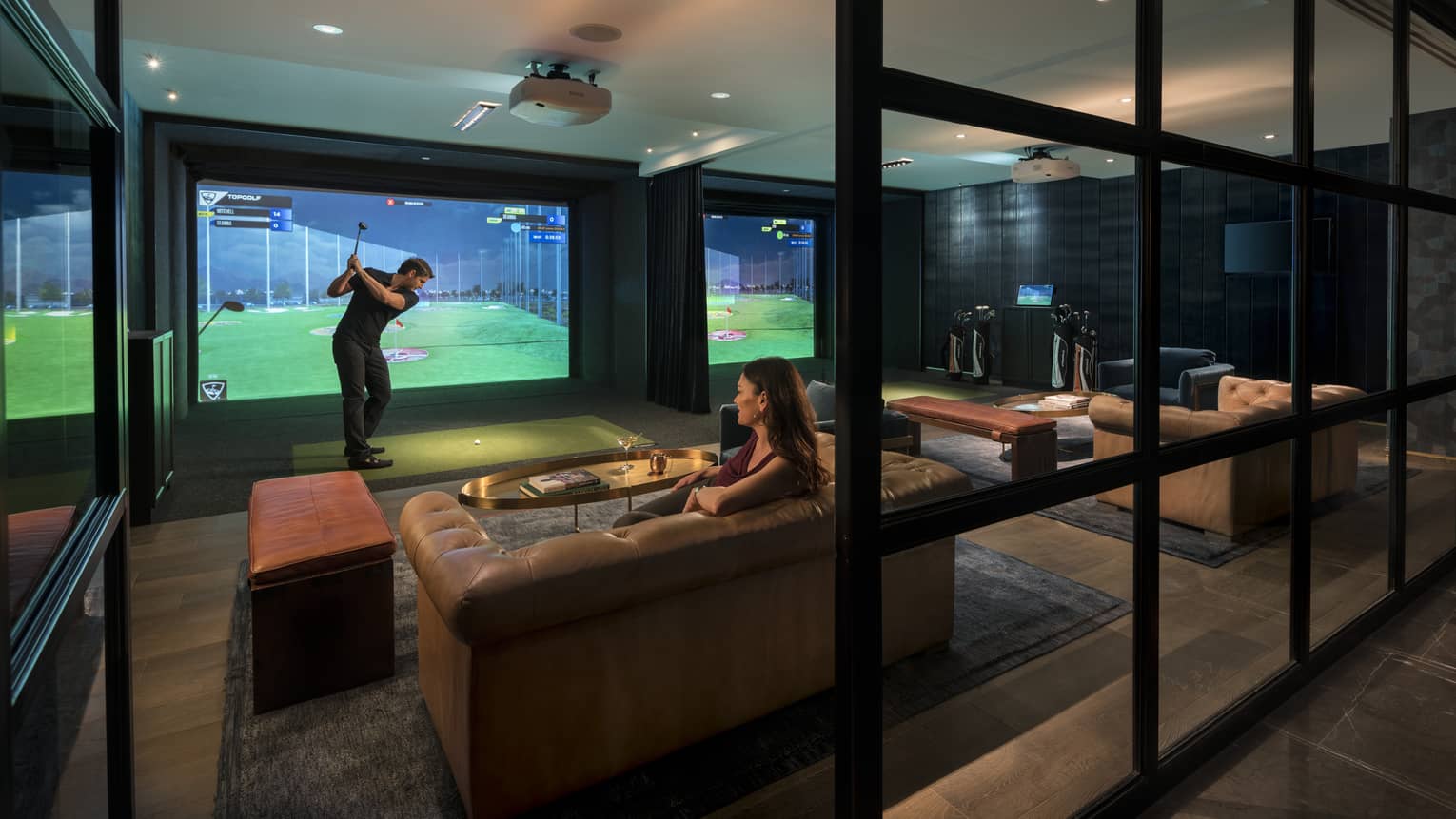 Topgolf lounge, man swings club by large golf simulator screen, woman sits on leather sofa behind him