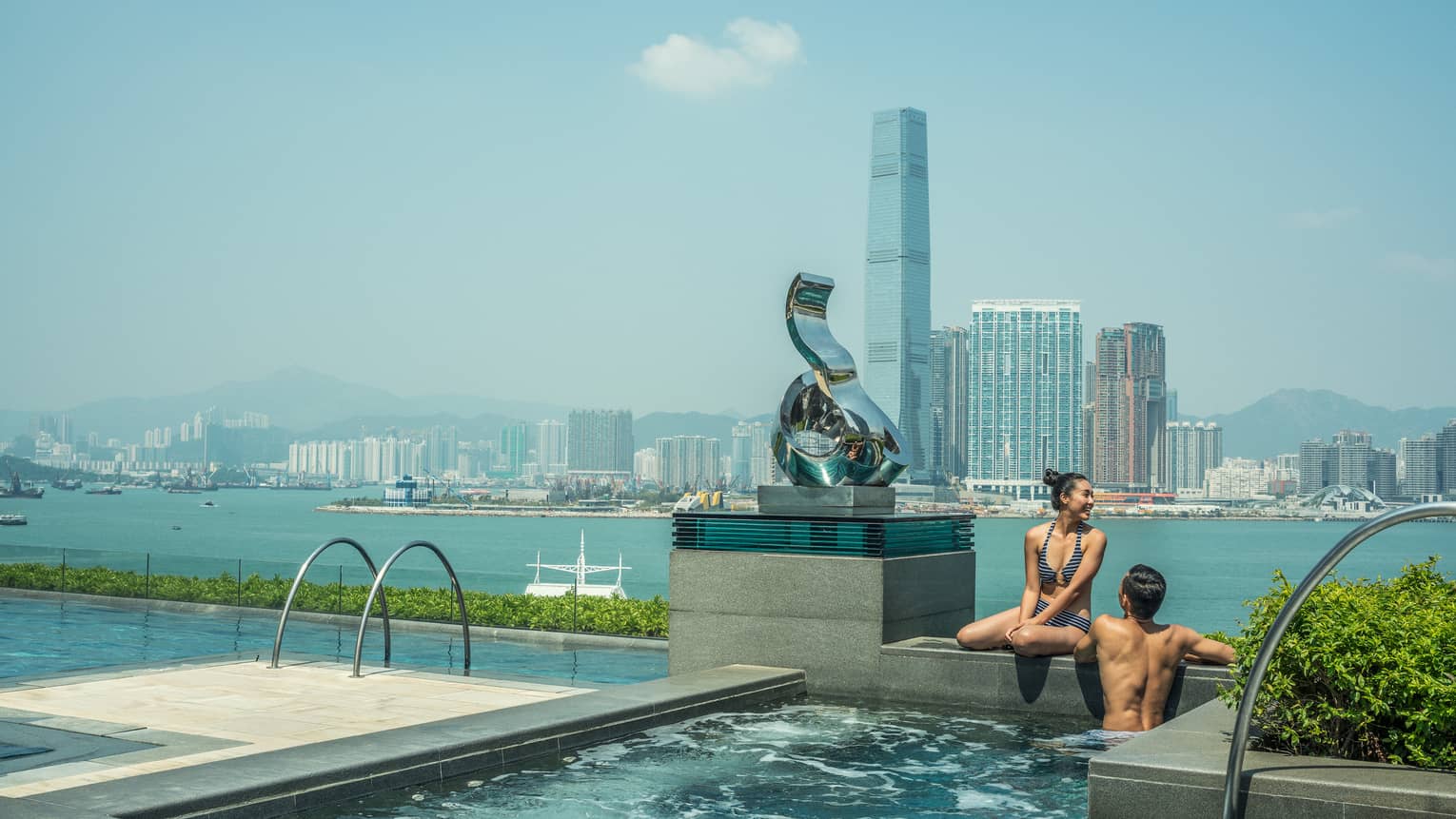 Couple wearing swimsuits wade at edge of outdoor swimming pool with sculpture, overlooking city