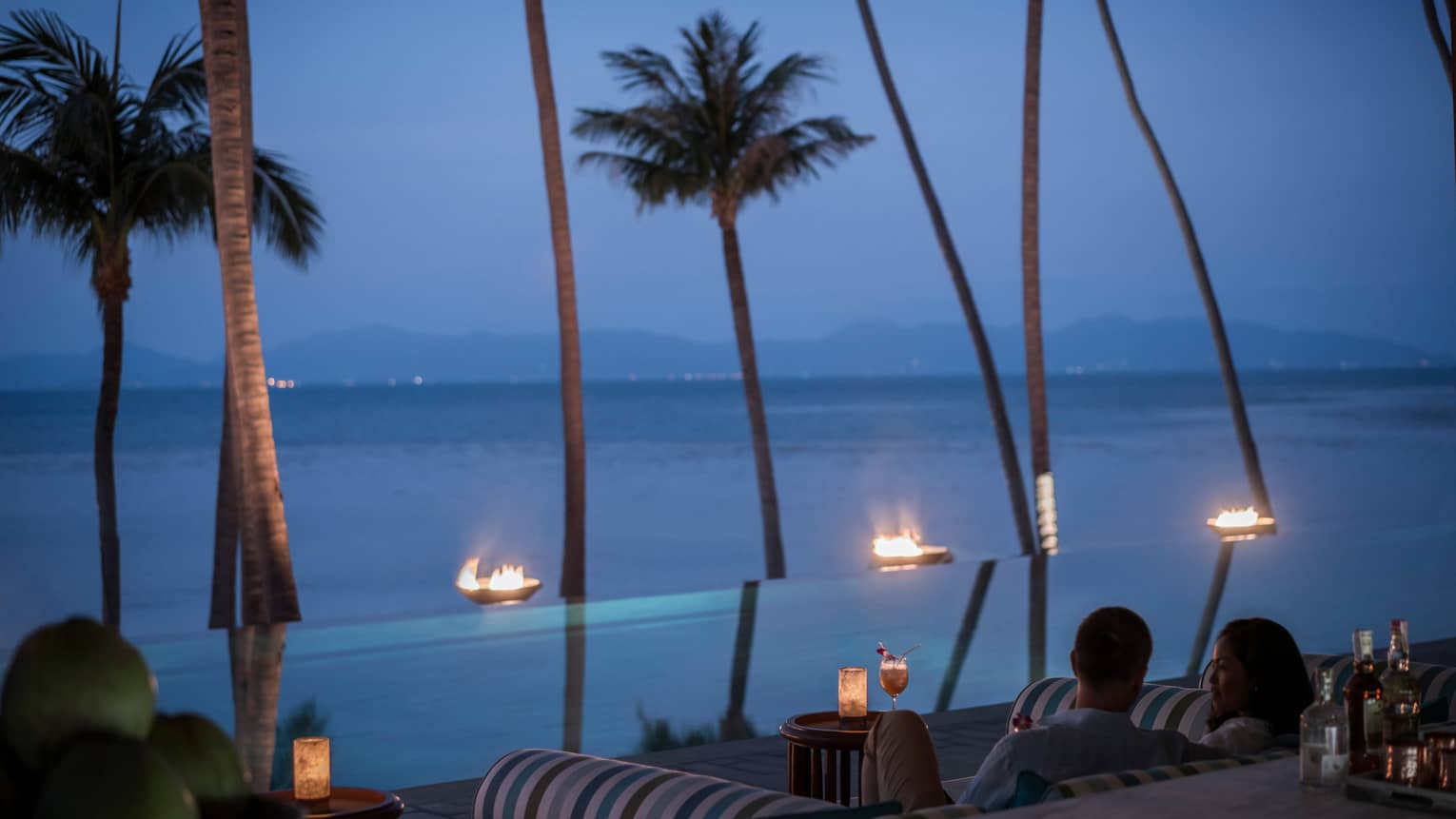 CoCoRum patrons look out at tall palm trees, outdoor fireplace line infinity pool in front of ocean at night