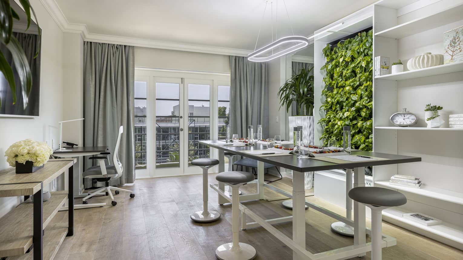A brightly lit office with a large table, stools, a desk, shelves, and plants on the wall.
