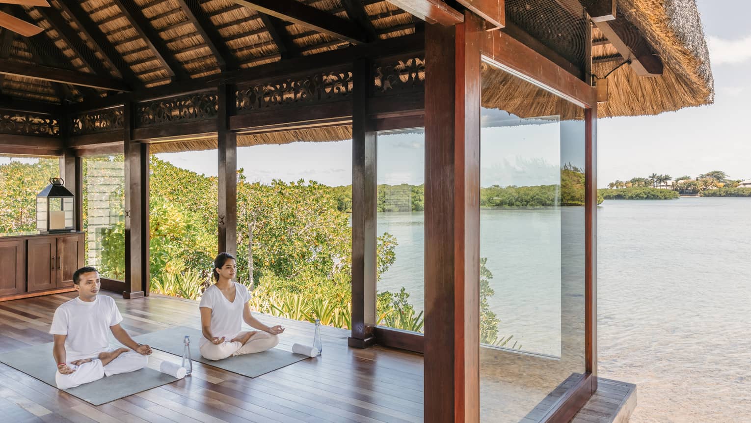 Two people wearing white seated in yoga poses in open-air spa yoga pavilion by beach