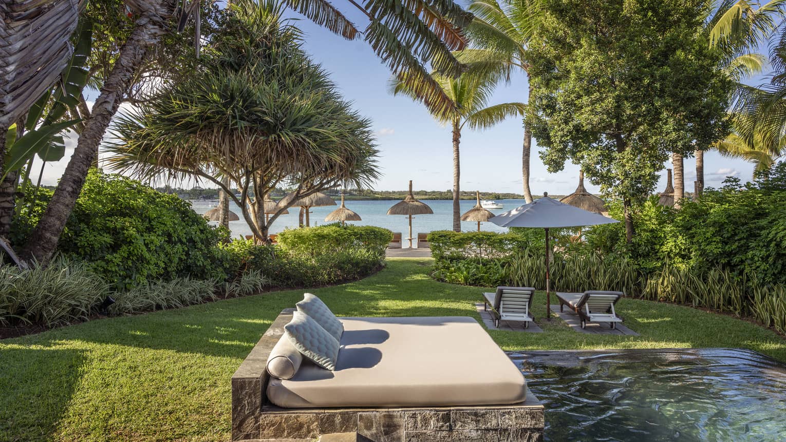 Tropical lawn with pool and day bed, two lounge chairs and umbrellas facing the beach