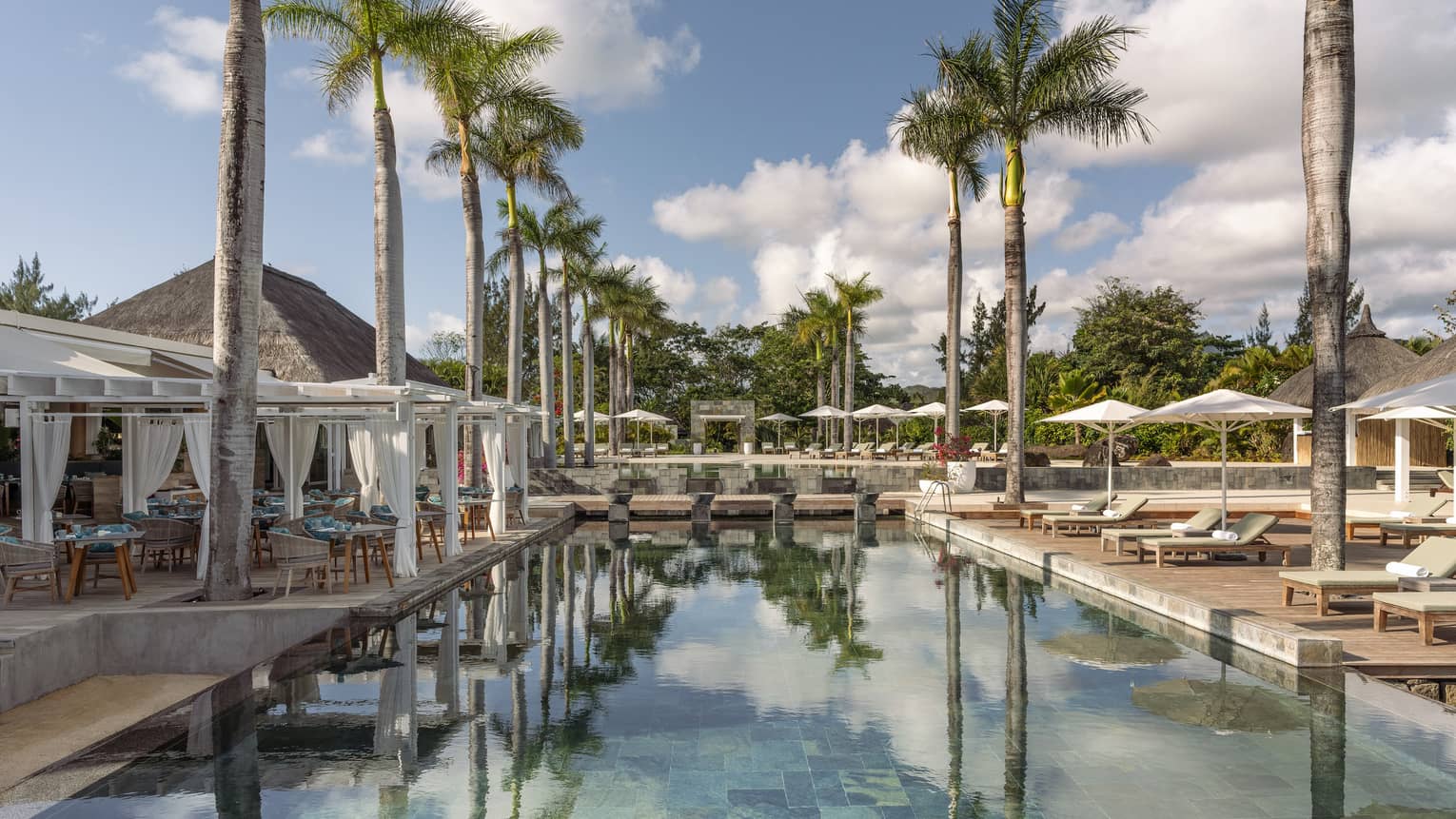 Outdoor pool flanked by palm trees, cabanas and lounge chairs with umbrellas
