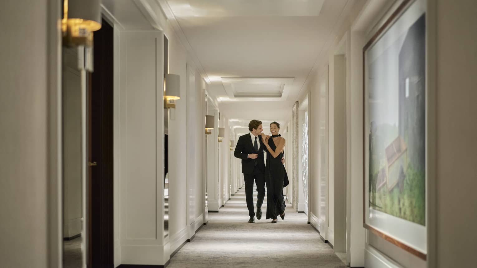 A couple elegantly dressed in formal attire walks arm in arm down a hotel hallway. The hallway is adorned with modern lighting and framed artwork, creating an upscale and sophisticated atmosphere.